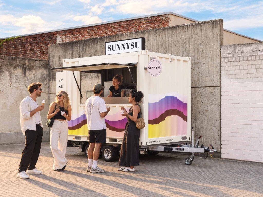 Sunnysu Softeis-Foodtrailer mit mehreren entspannten Gästen an einem sonnigen Abend – mobile Walkingbox im urbanen Innenhof mit Frozen Yogurt und Softeis.